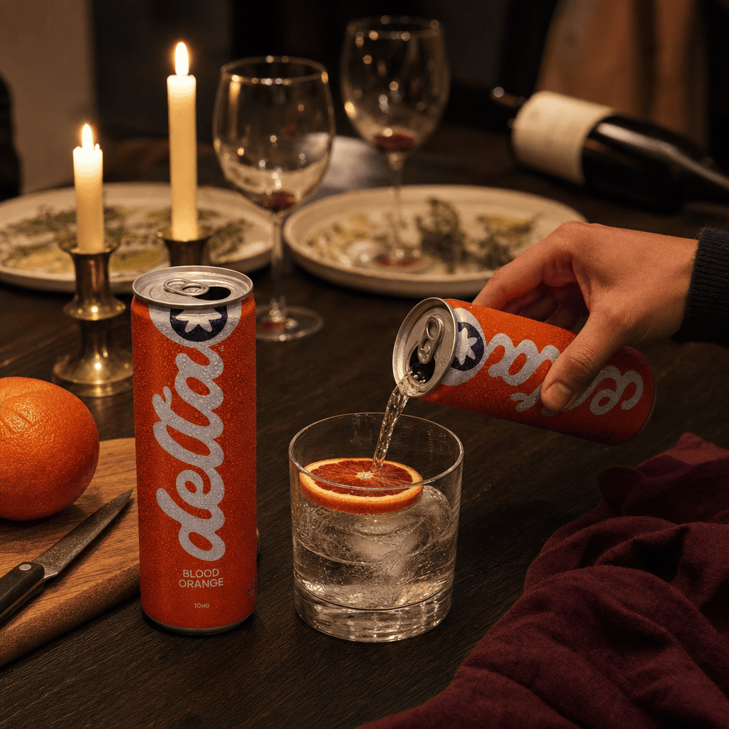 Hand pouring a Delta Blood Orange can into a clean rocks glass with ice and a fresh blood orange slice, candlelit dinner table with the room's emptied wine glasses behind