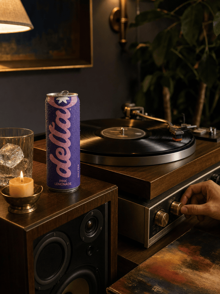 Vinyl record spinning on a wood-and-brass turntable, a Bold Pink Lemonade can on the speaker, dim warm lamp, hand reaching for the volume knob
