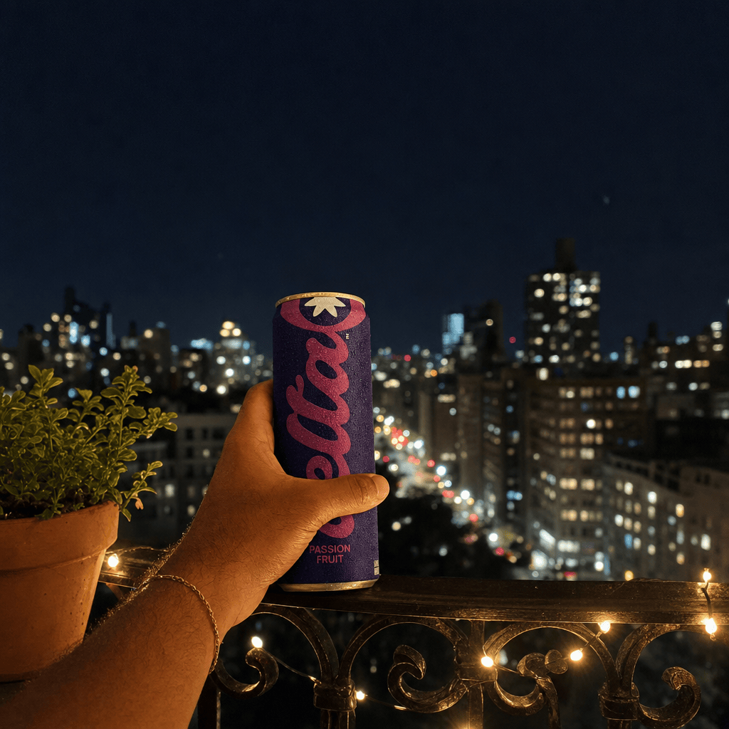 A hand on a black wrought-iron balcony rail at night holding a Bold Passion Fruit can, blurred city lights below