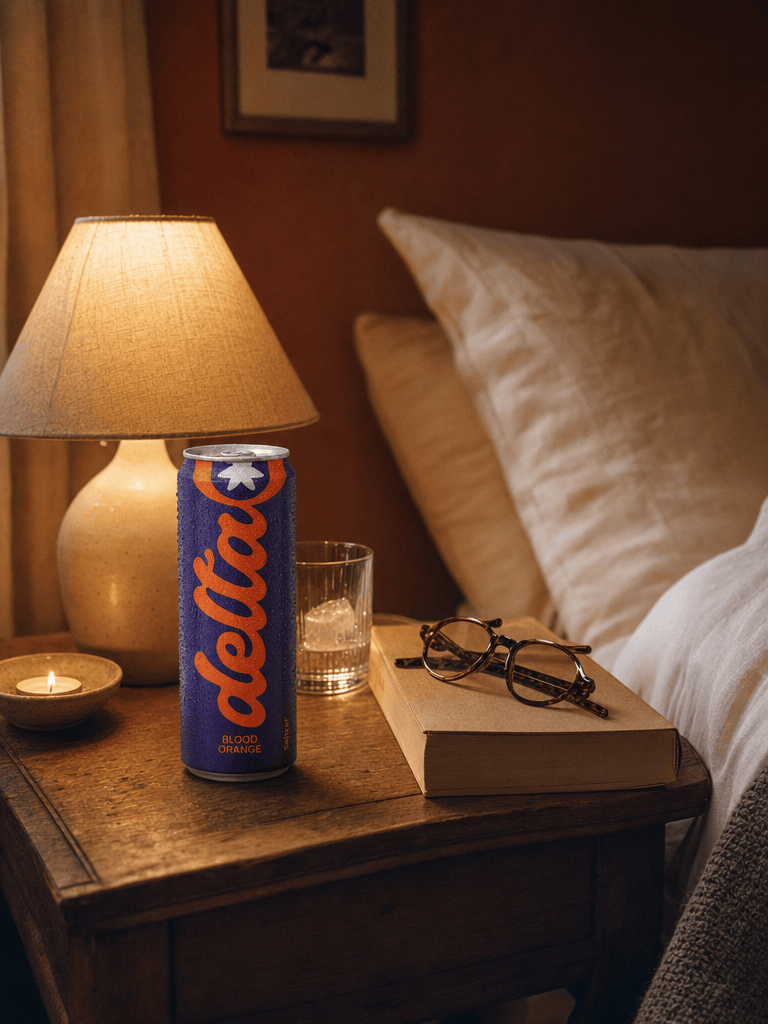 A bedside table at night with a Bold Blood Orange can, paperback open face-down, glasses, dim warm bedside lamp